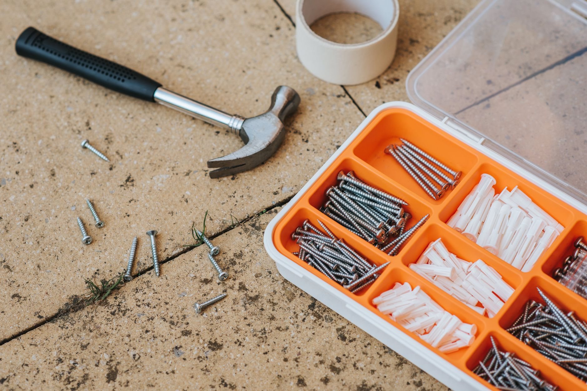 Box of assorted repair instruments and screws next to a hammer on a tile floor
